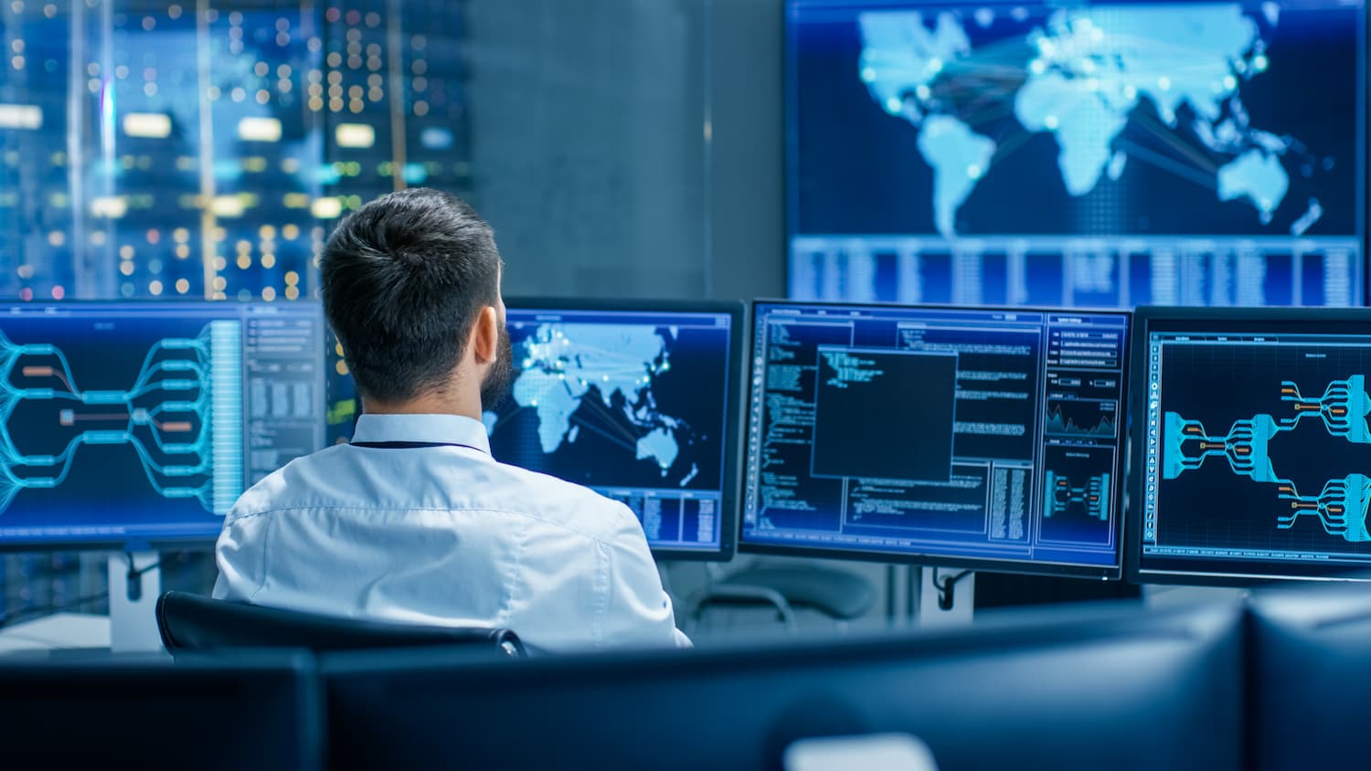 Hombre sentado frente a tres pantallas de computadora en un centro de datos, trabajando en múltiples tareas tecnológicas.