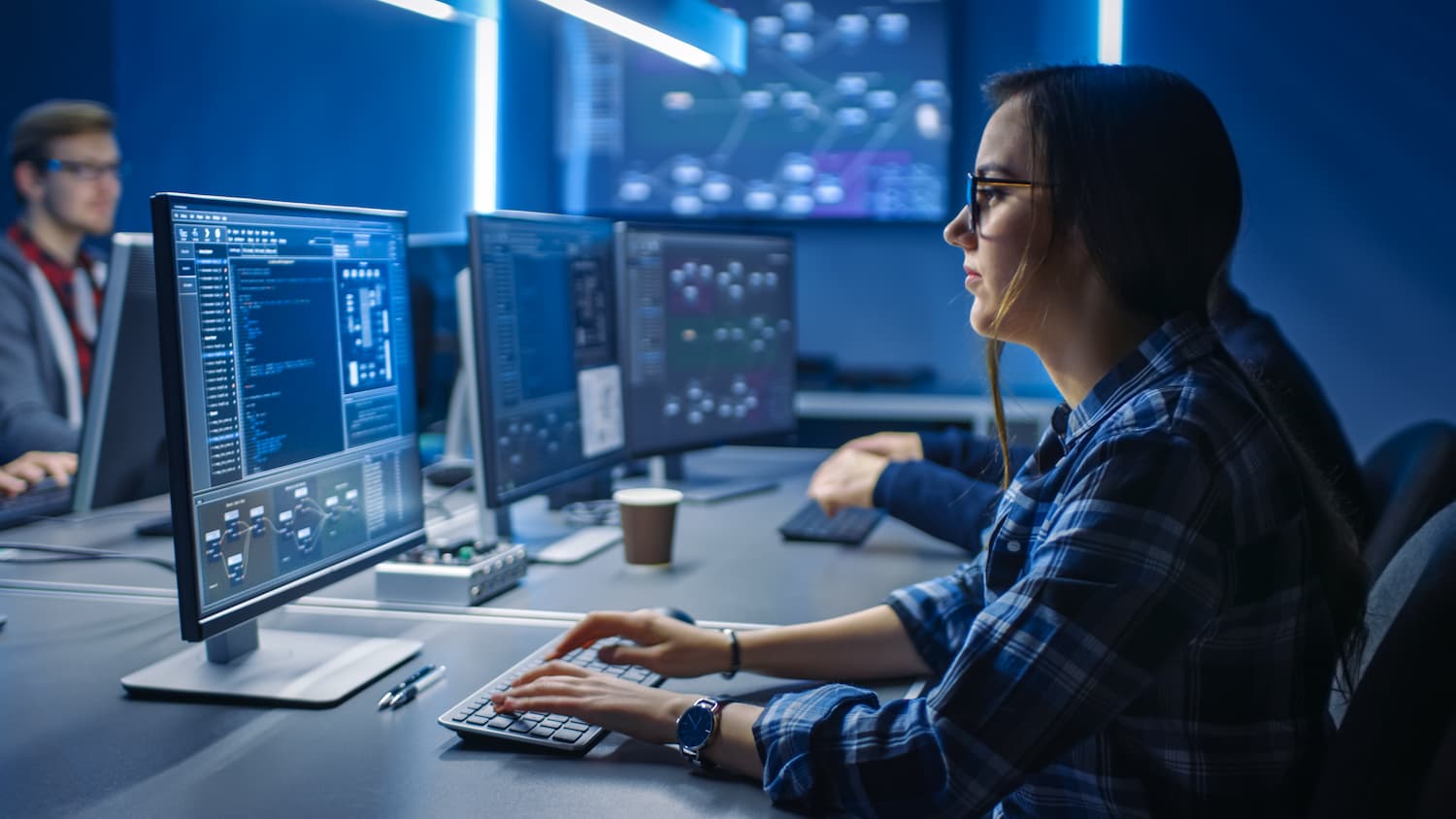 Mujer trabajando en una computadora en una sala oscura de un centro de datos, rodeada de tecnología avanzada.