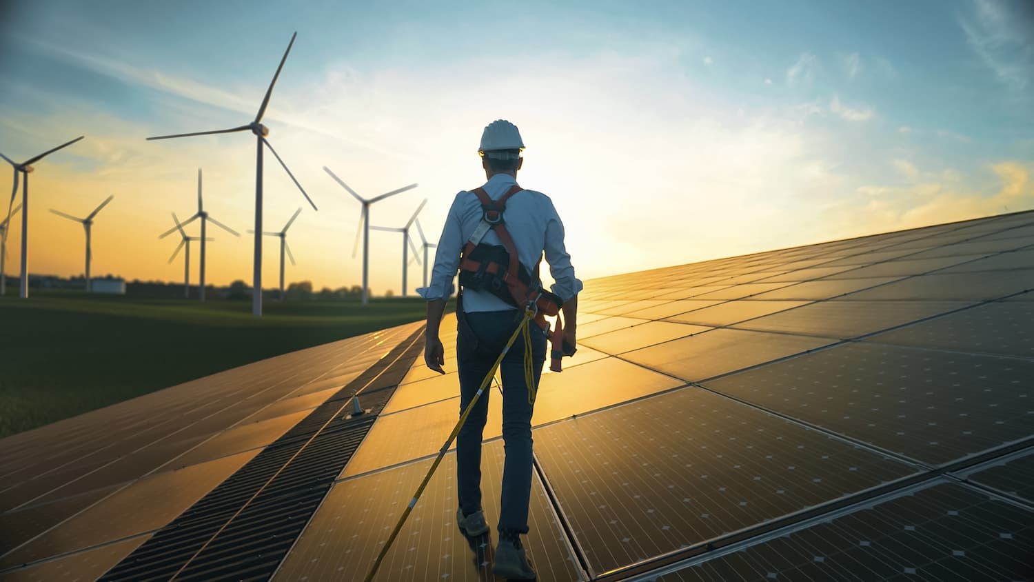 Hombre de pie sobre un panel solar, con turbinas eólicas al fondo, simbolizando la tecnología sostenible.